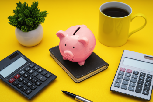 A piggy bank on a notebook surrounded by calculators, a plant and a yellow coffee cup