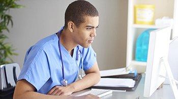 Young male presenting doctor wearing scrubs looking at a computer.