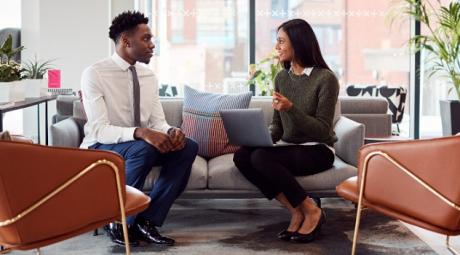 Two individuals sitting on a couch facing toward each other and speaking, as if they are in the middle of an interview.