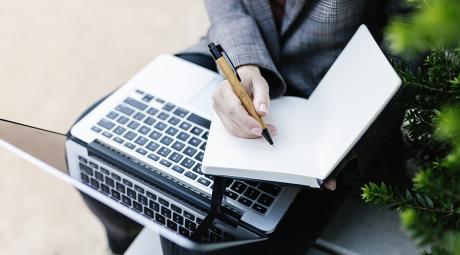 Close-up of a hand writing in a notebook next to a laptop