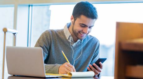 A student looks at a phone while taking notes next to a laptop