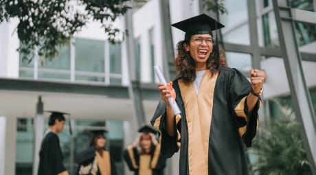 A graduate in a cap and gown holding a diploma celebrates