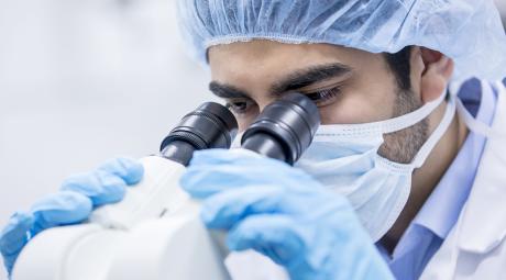 A researcher wearing a mask looks through a microscope
