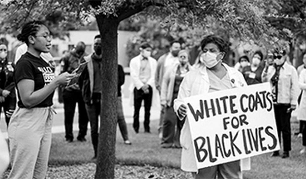 Group of people participating in an anti-racism protest. One of them is holding a "White Coats for Black Lives" sign.