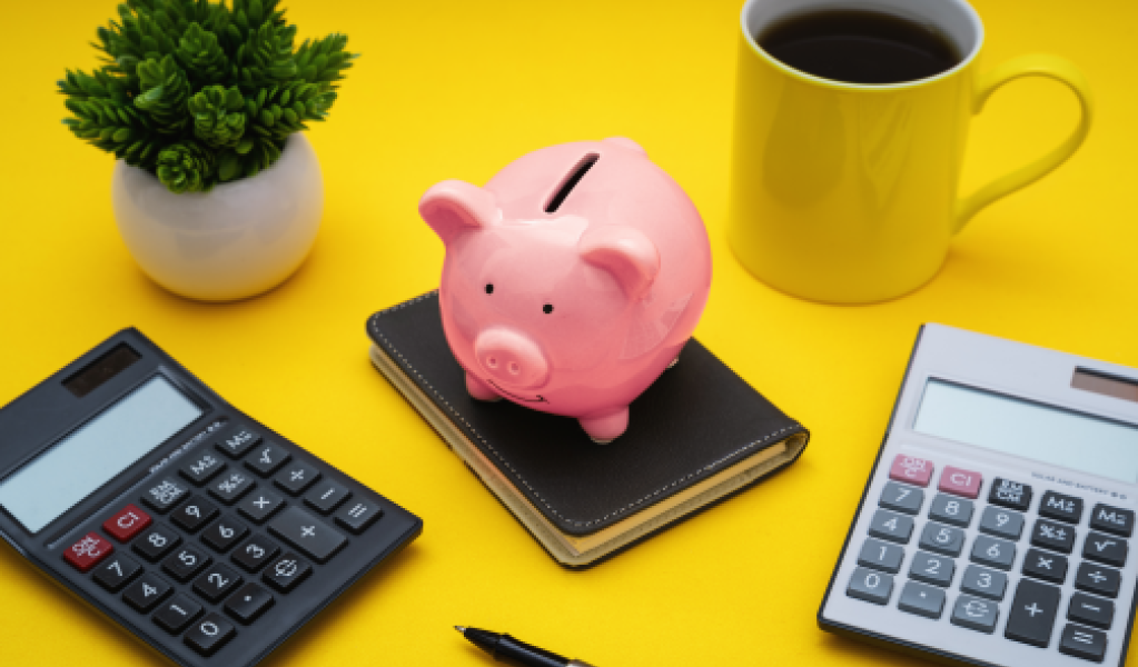 A piggy bank on a notebook surrounded by calculators, a plant and a yellow coffee cup