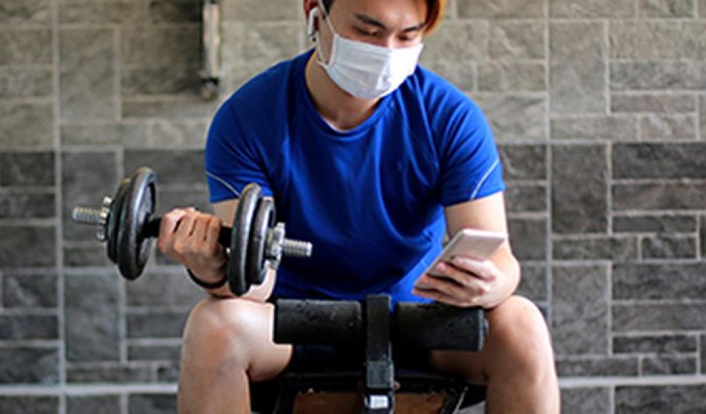 A person wearing a mask uses a phone while lifting weights