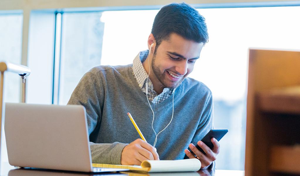 A student looks at a phone while taking notes next to a laptop